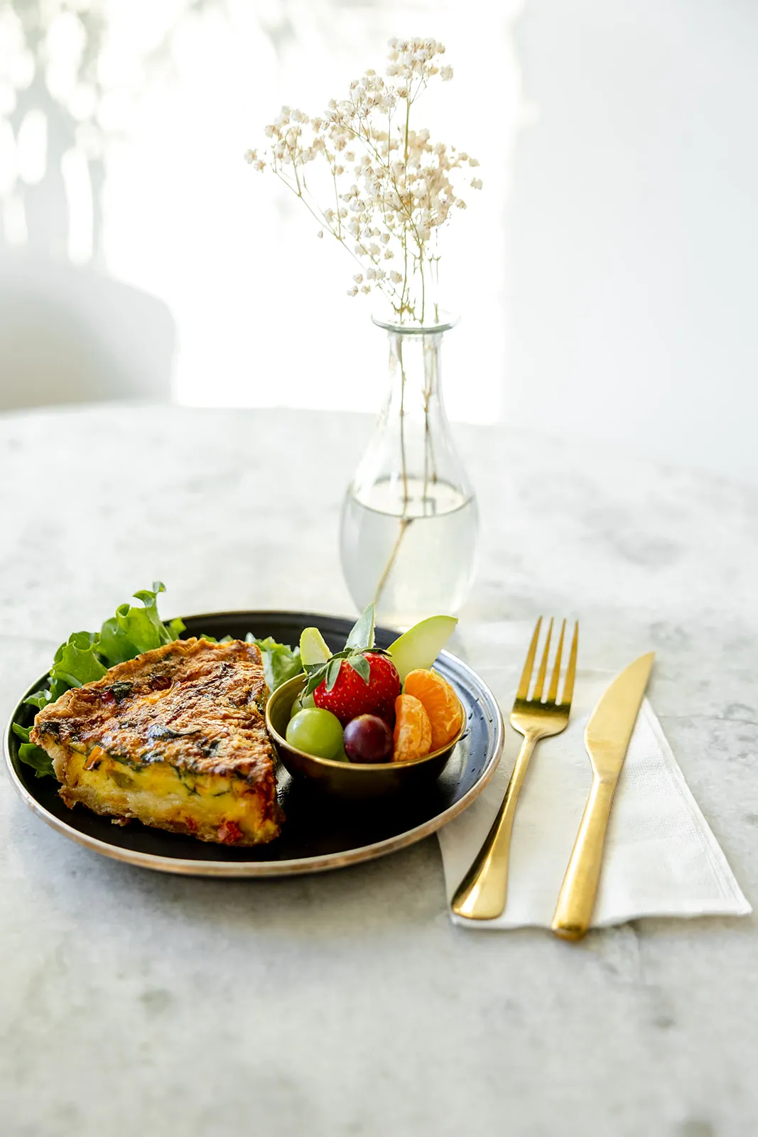 House-made quiche with side fruit cup, plated on marble with gold flatware